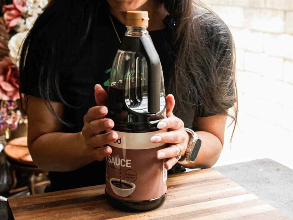 A person with long hair holds a large bottle of soy sauce on a wooden surface. Only their hands and torso are visible. Light comes in from a window in the background.